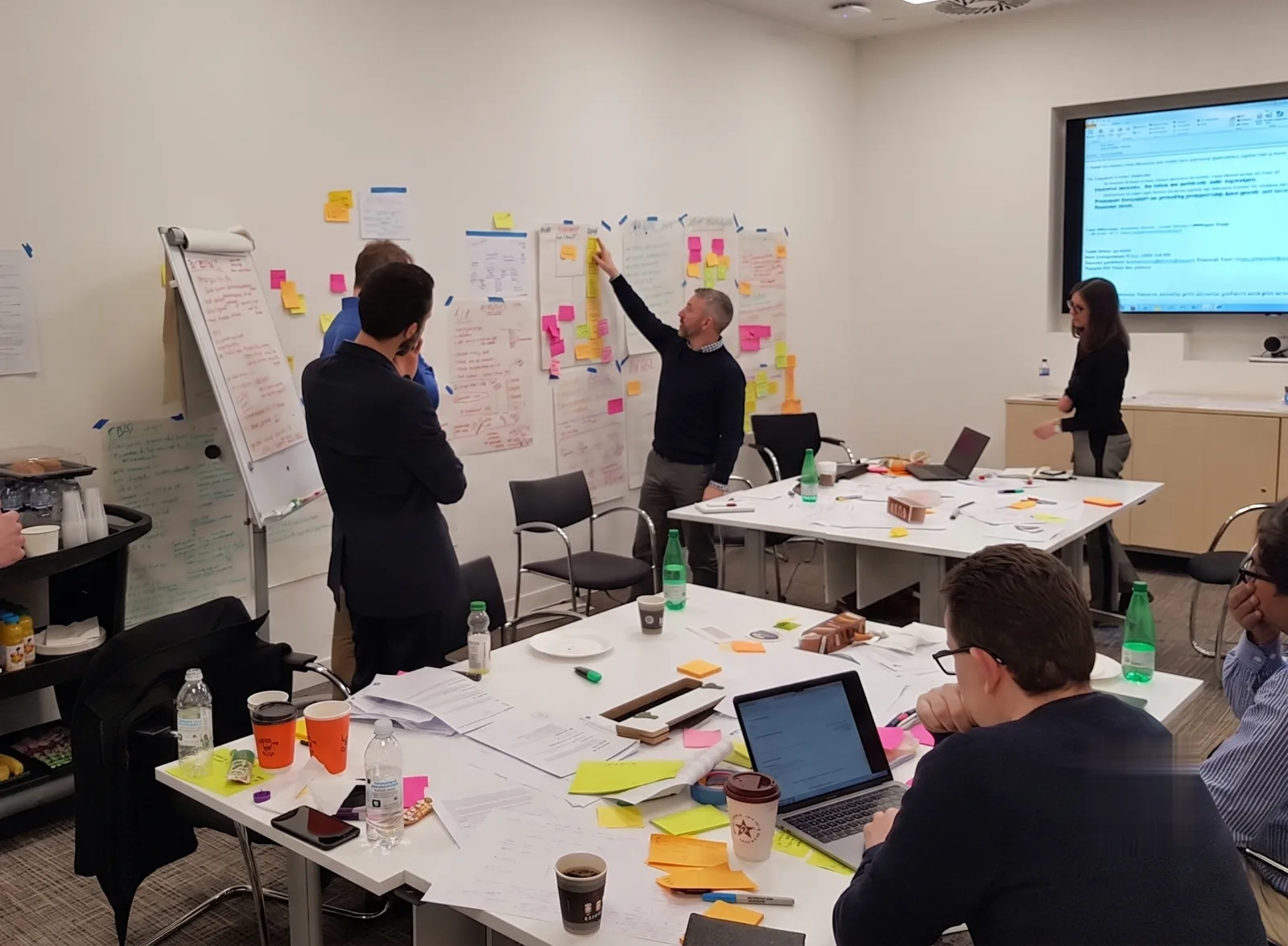 A working session in progress: teams mapping ideas on a wall covered in sticky notes and flip-chart sheets